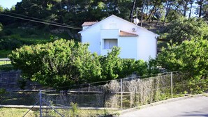 A white single-story house with a tiled roof, surrounded by dense green trees and a chain-link fence. - Apartment Ribaric (Rab)