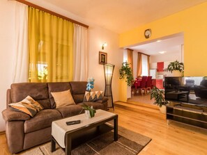 Un salon avec un canapé marron, une table basse et un meuble pour la télévision. On aperçoit à l’arrière-plan un coin repas avec une table et des chaises rouges.