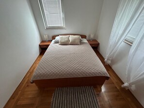 A neatly made bed with a quilted cover, wooden nightstands with lamps, a window with blinds, and a striped rug on a wooden floor. - Apartment Pogi Srima (Vodice)