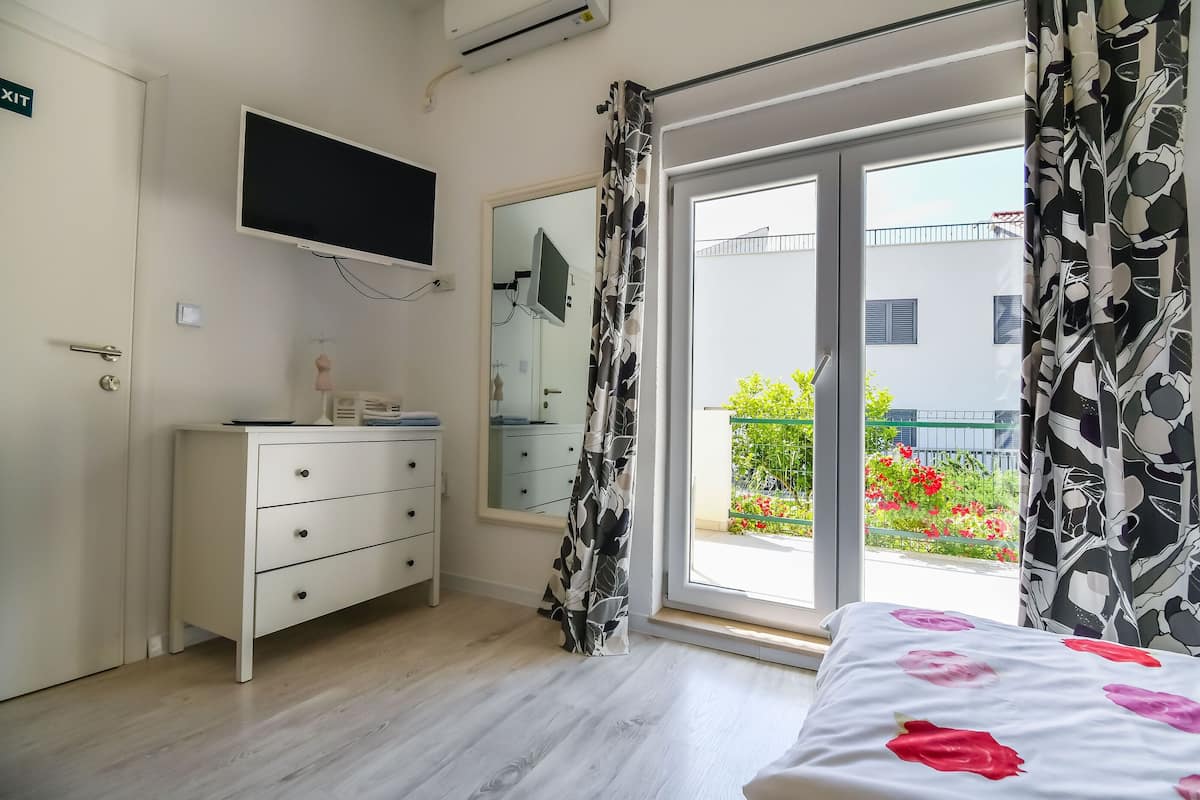 Un dormitorio con cama, cómoda, televisor montado en la pared, ventana con vista a un balcón y una puerta señalizada con 'EXIT'.