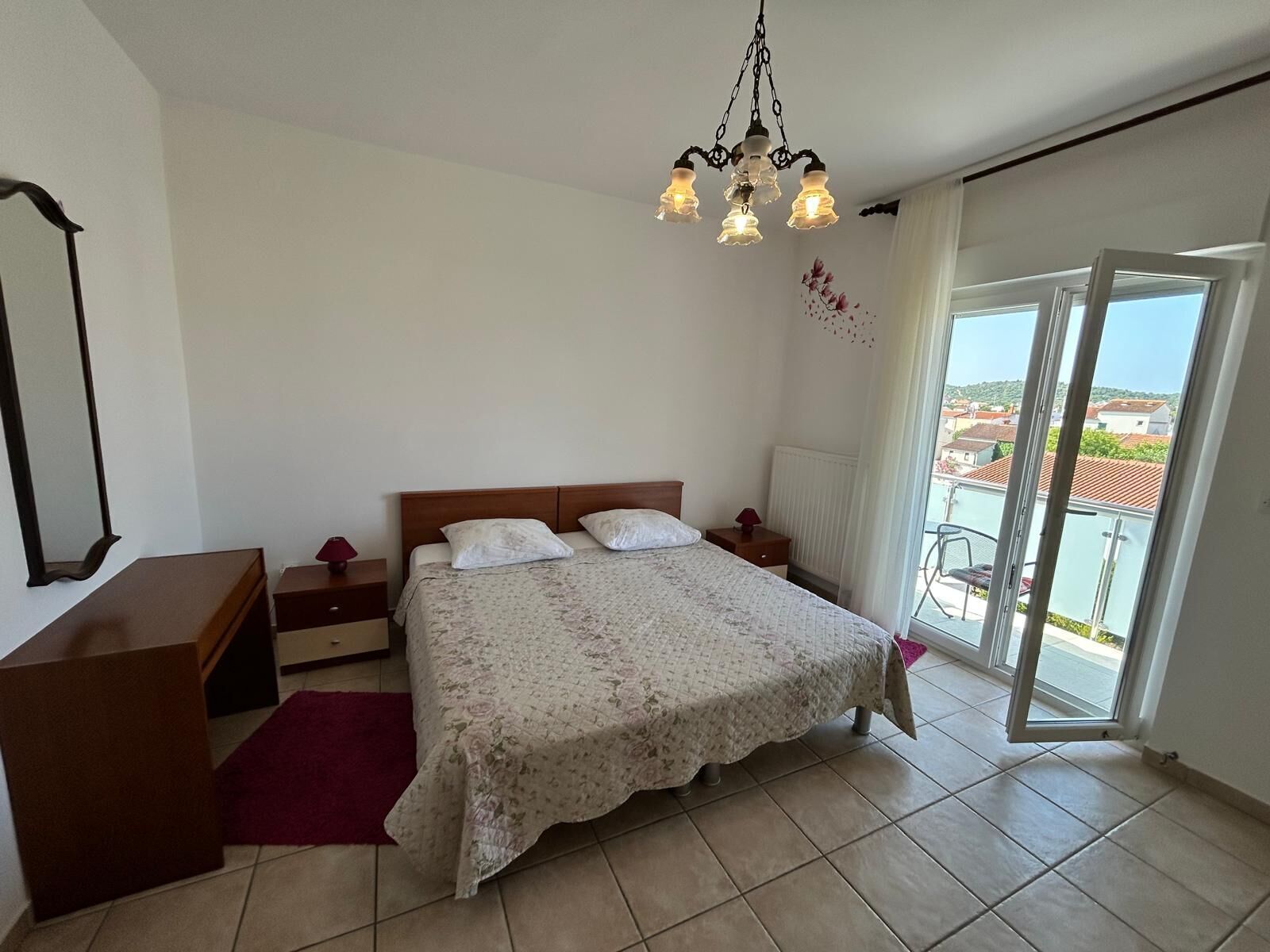 A bedroom with a bed, bedside tables, a dresser, a chandelier, and a balcony with a view.