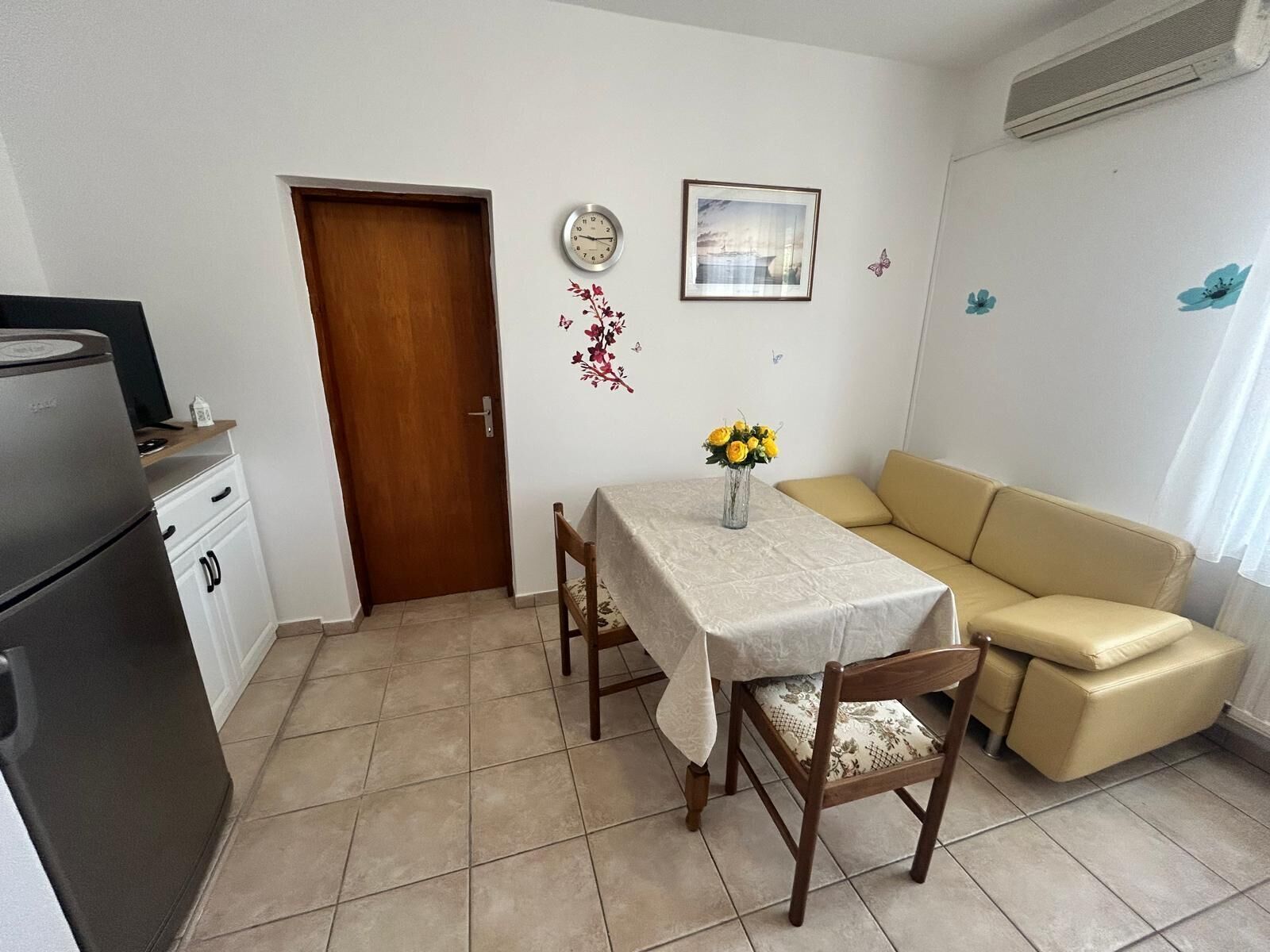 A small, clean room with a dining table, a sofa, a refrigerator, and a door.