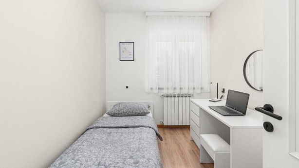A small, well-lit bedroom with a single bed, a desk with a laptop, a radiator, and a window with sheer curtains. - Apartment Vista Olivo (Brtonigla)