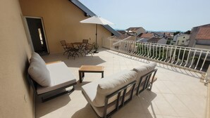 Un balcon avec un canapé, une petite table et une chaise. Une porte donne accès à l’intérieur, et une balustrade longe le bord.
