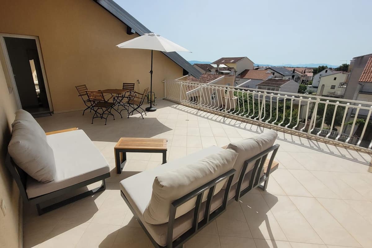 A balcony with a sofa, a small table, and a chair. There is a door leading to the interior and a railing along the edge.