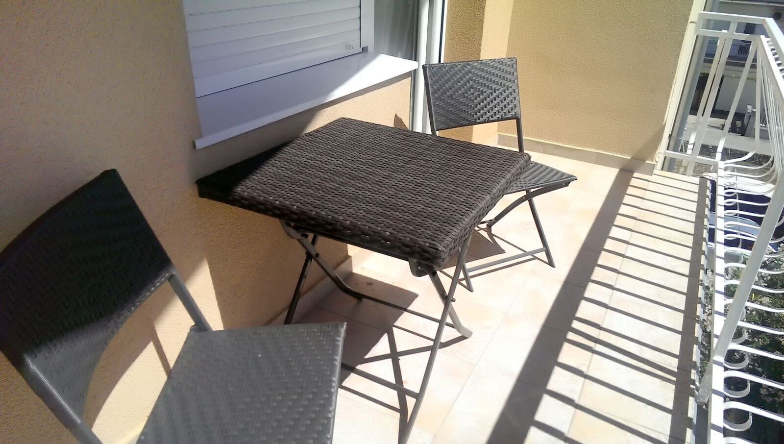 Uma pequena varanda com mesa e duas cadeiras, uma janela e grade de metal.