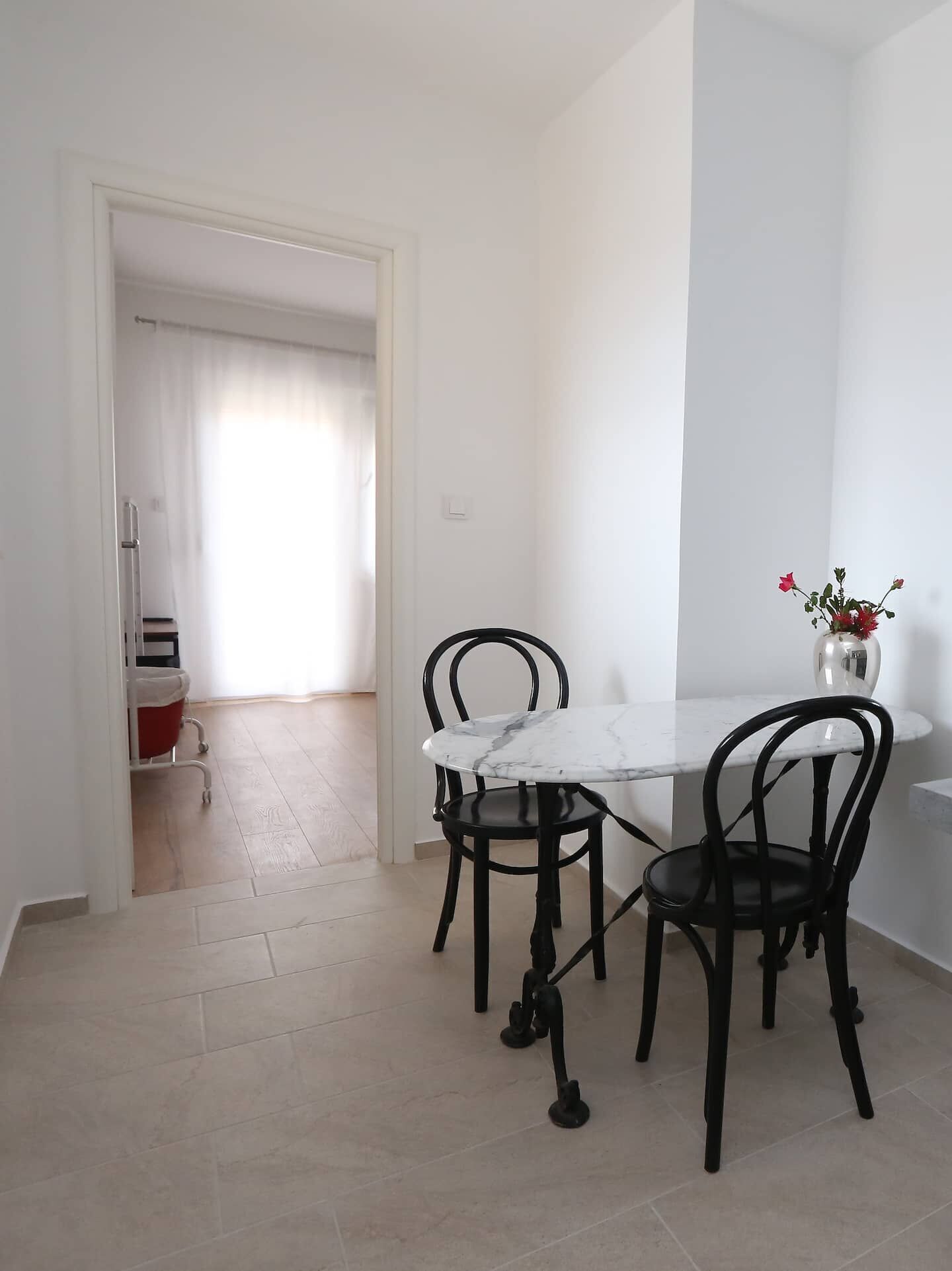 A dining area with a white marble table and black chairs, a vase with flowers on the table, and a view of a kitchen area with a refrigerator and a red chair.