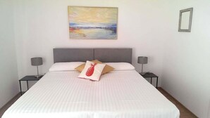 Un lit bien fait, recouvert d’une courtepointe blanche et agrémenté de deux oreillers, dont l’un est orné d’un motif ananas, dans une chambre dont la tête de lit est grise.