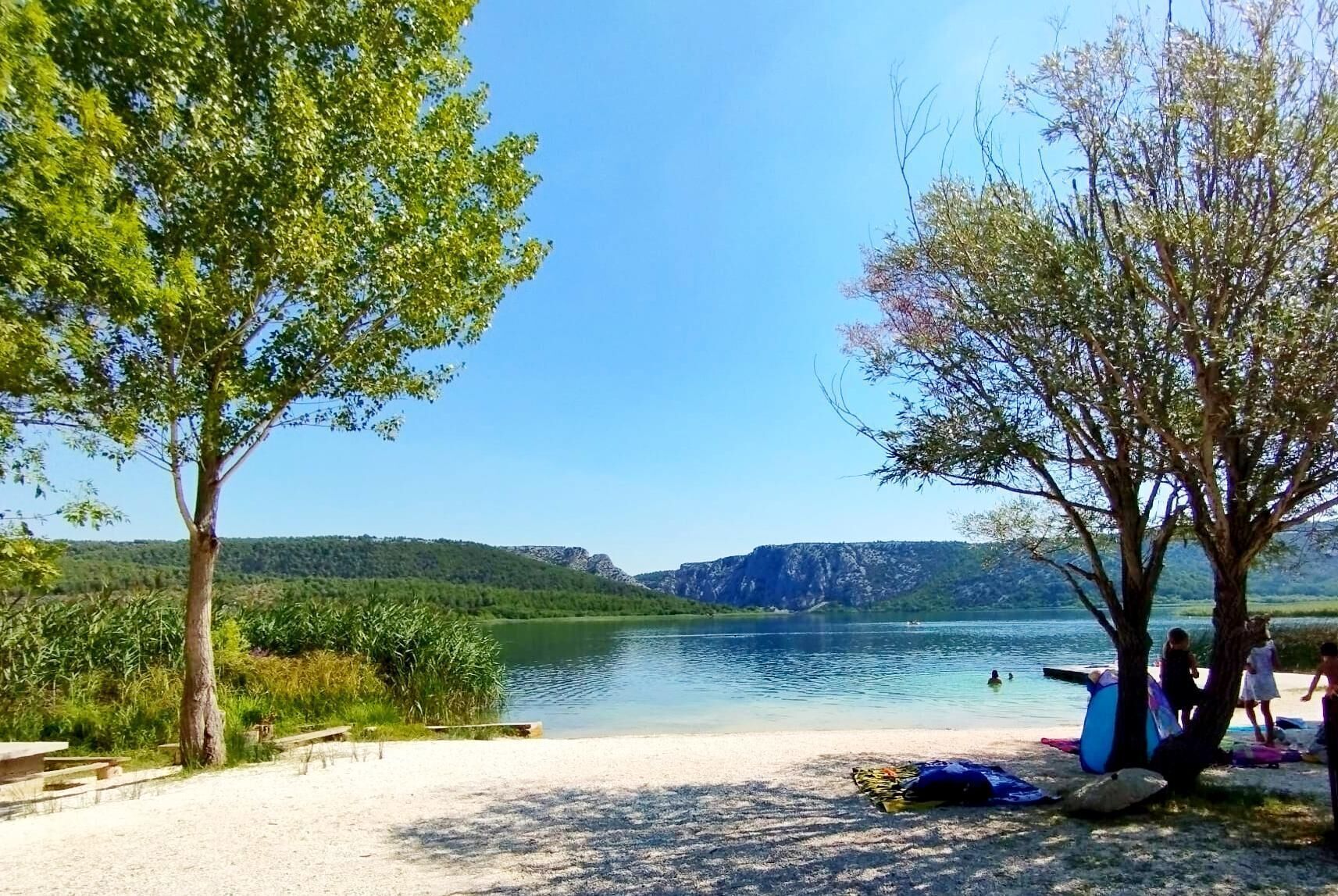 Ein See mit Strand, Bäumen, sandigem Ufer und freiem Blick auf den See und ferne Berge.