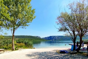 Una playa junto al lago, con árboles, orilla arenosa y una vista despejada del lago y montañas distantes.
