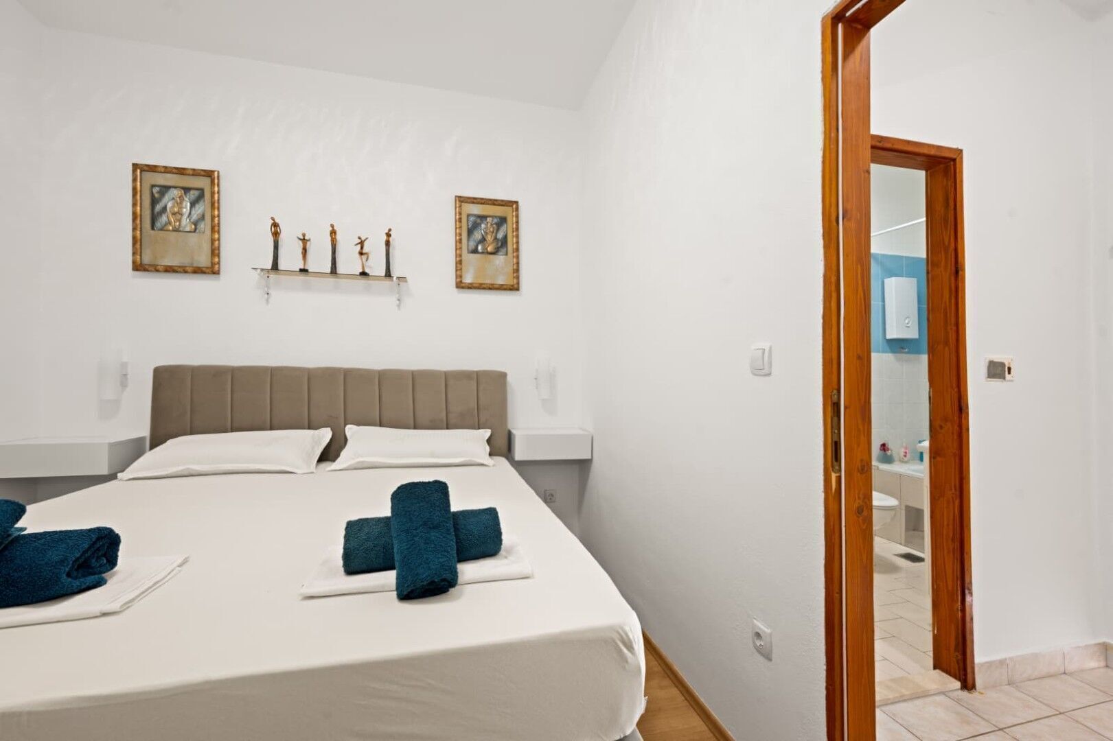 A hotel room with a large bed, two wall-mounted picture frames, a wooden door, and a small bathroom visible through the door.