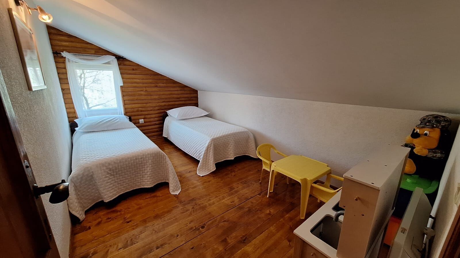 A small, cozy room with two single beds, a wooden ceiling, a small yellow table, and a toy figure.