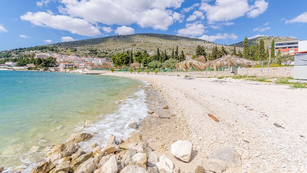 Una spiaggia con acque cristalline turchesi, un litorale roccioso e sullo sfondo un paesaggio collinare con edifici e alberi.