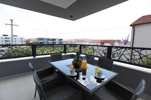 A balcony with a table set for breakfast, including fruits and beverages, overlooking a residential area with buildings and a green field. - Apartment Ivana (Trogir)