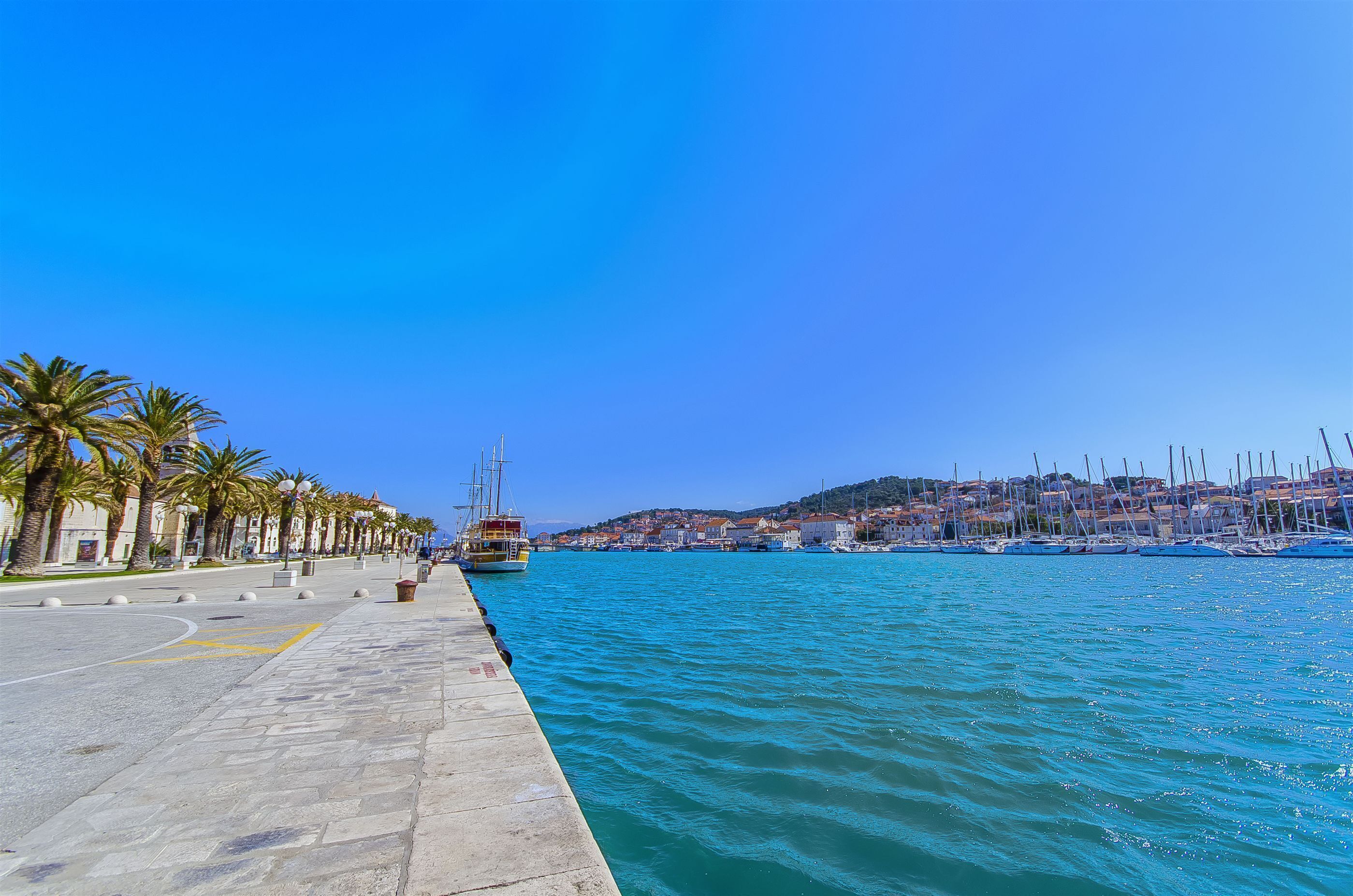 A waterfront promenade with a row of palm trees, a dock with a sailboat, and a cityscape in the background.