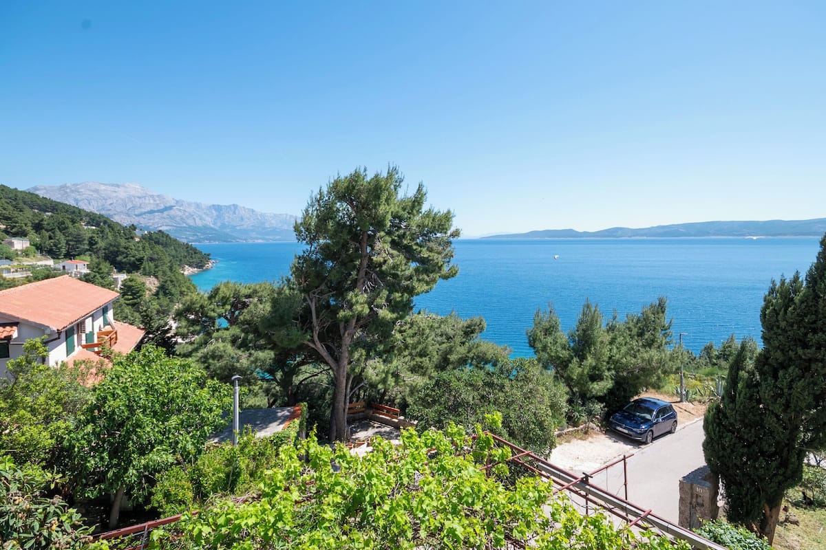 A coastal view with a clear blue sea, a mountain range in the background, and a residential area with houses and greenery.