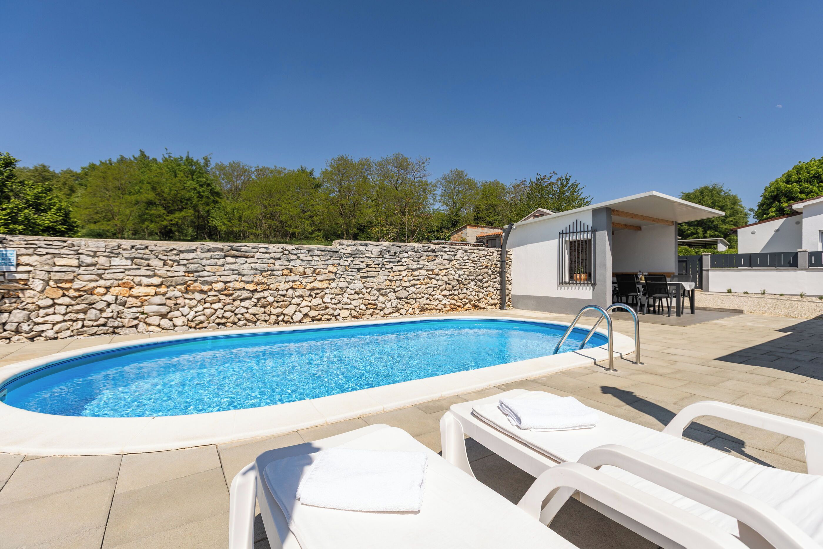 A swimming pool with lounge chairs, a stone wall, and a small building with a patio.