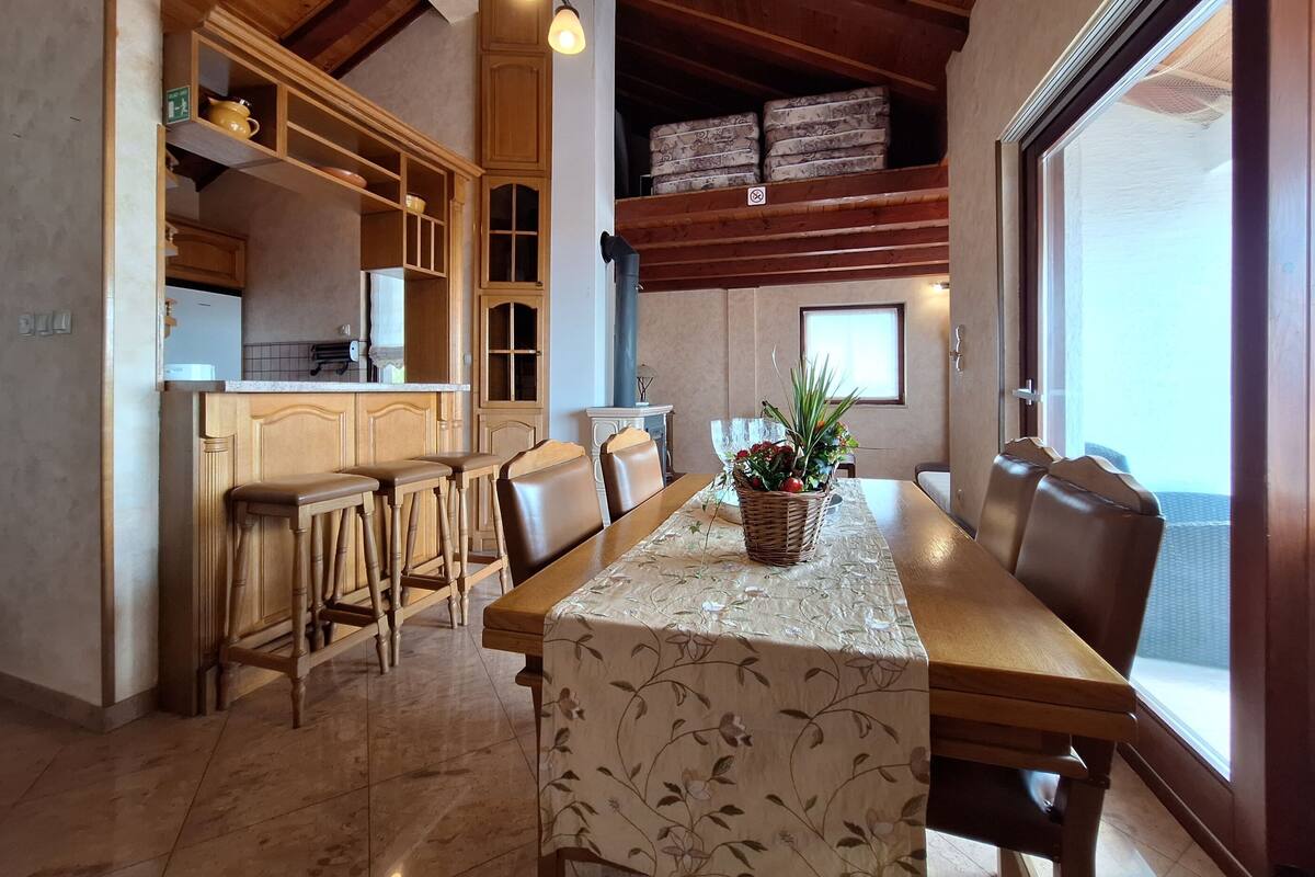 A dining area with a wooden table, chairs, and a floral tablecloth. There is a kitchen area with wooden cabinets and a refrigerator.