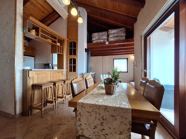 A dining area with a wooden table, chairs, and a floral tablecloth. There is a kitchen area with wooden cabinets and a refrigerator. - Apartments Preden (Crikvenica)