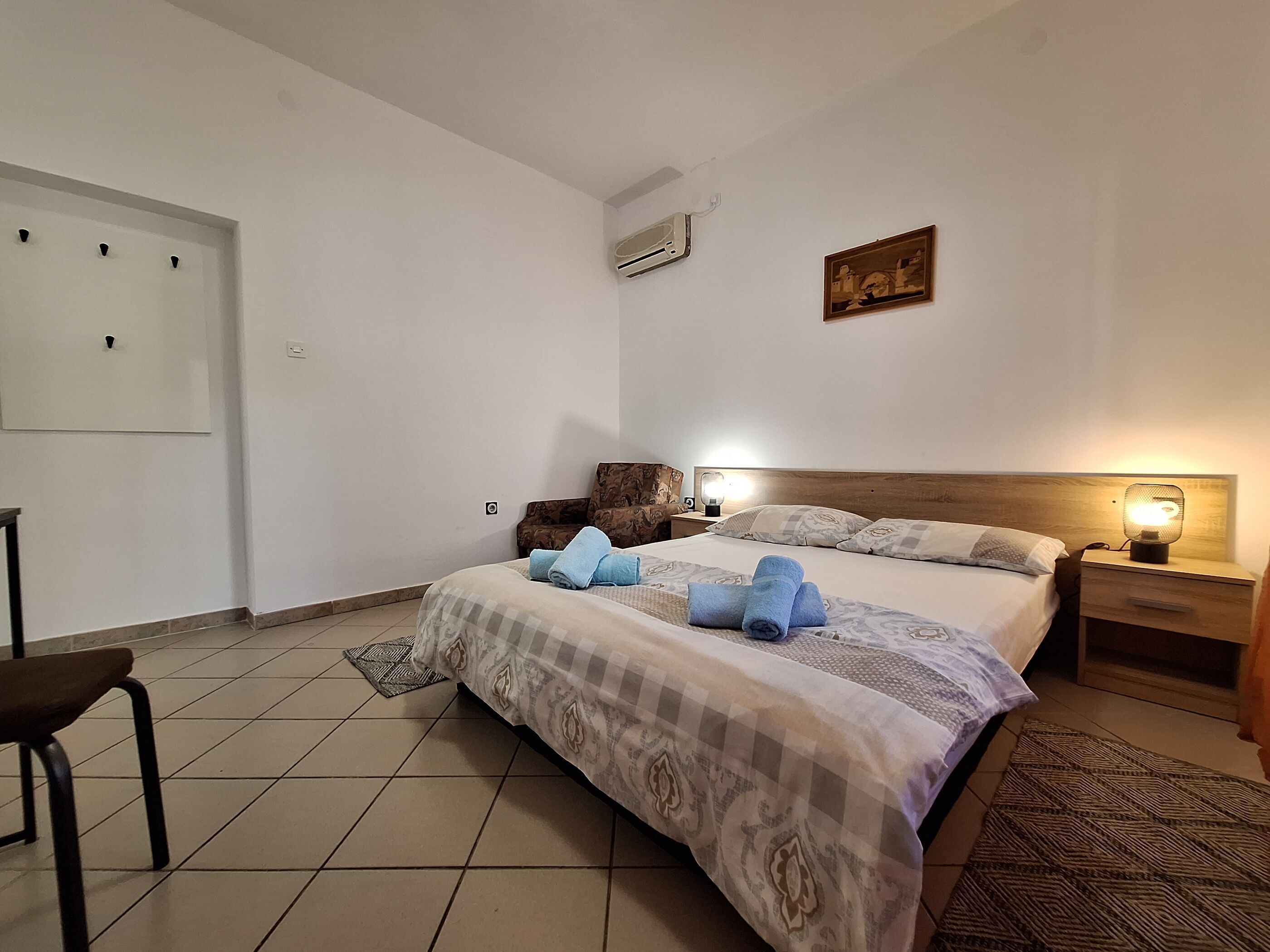 A hotel room with a bed, a nightstand, a chair, a wall-mounted air conditioner, and a painting.