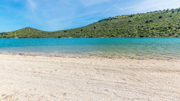 Uma praia com águas turquesa cristalinas, uma faixa de areia e uma colina verde ao fundo.