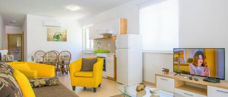 A modern living room with a sofa, yellow armchair, and a glass coffee table. A kitchen with white cabinets and a refrigerator is visible in the background.