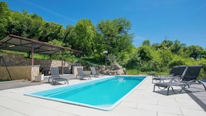 Una piscina rectangular con solárium empedrado, rodeada de sillas de descanso y una zona de estar cubierta.