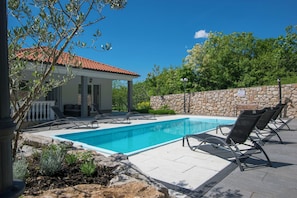 A swimming pool with lounge chairs, a stone wall, and a building with a red-tiled roof.