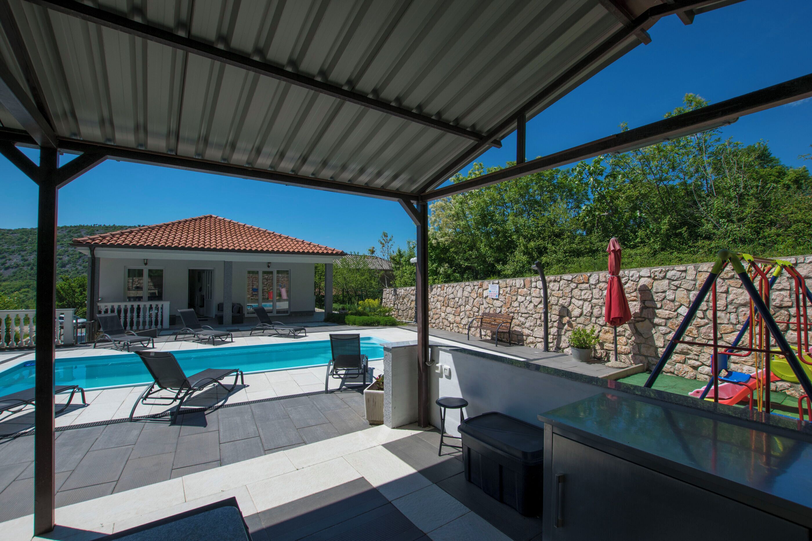 A covered outdoor area with a swimming pool, lounge chairs, and a stone wall with a playground.
