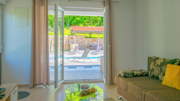 Una sala de estar con un sofá, una mesa de centro con objetos decorativos y vistas a un área de piscina exterior con sillones de descanso.