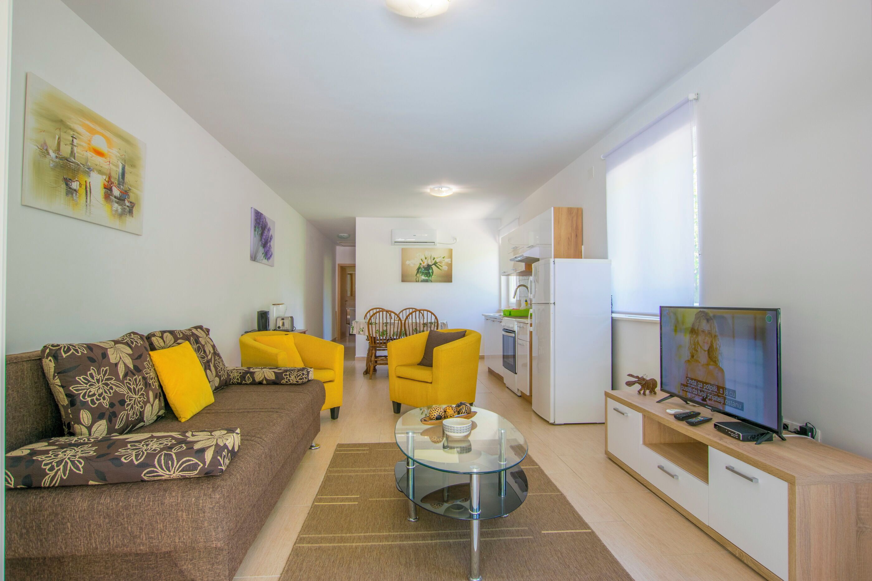 Un salon moderne avec un canapé, deux fauteuils jaunes, une table basse en verre et un téléviseur posé sur un meuble en bois.