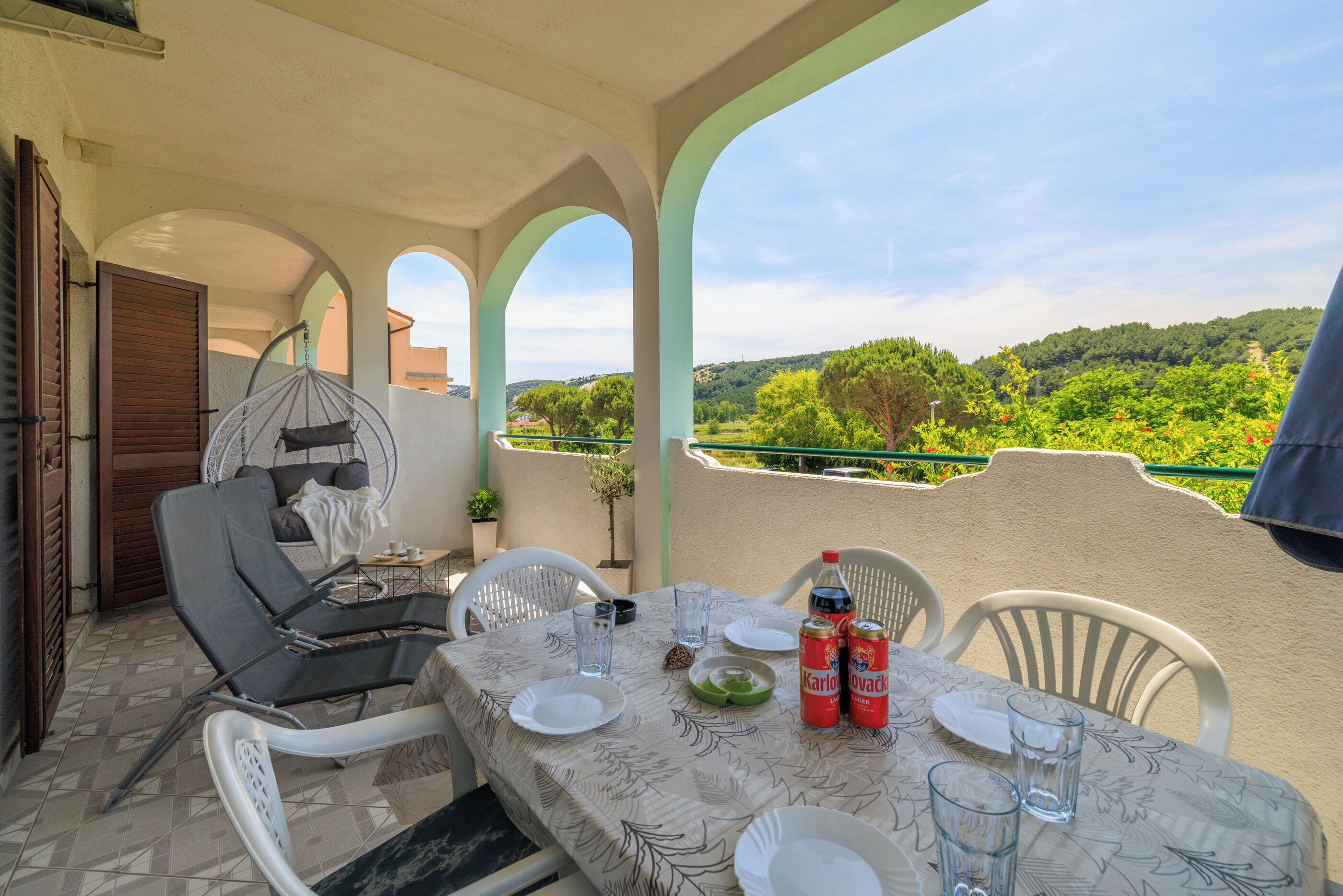 A terrace with a table set for dining, white chairs, a hanging chair, and a view of green hills.