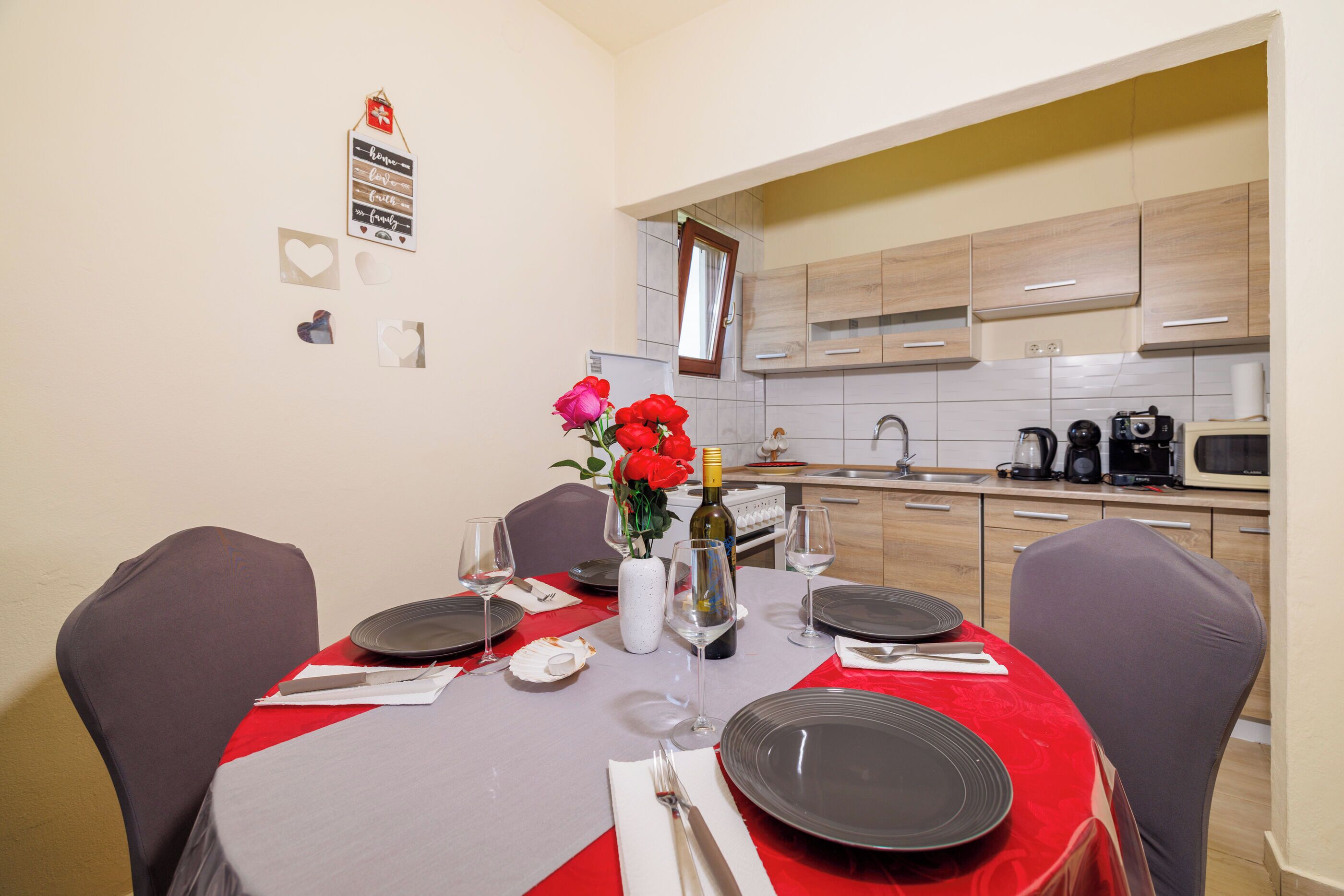 Een eetruimte met een tafel voor vier personen, een vaas met rode bloemen en een keuken op de achtergrond.