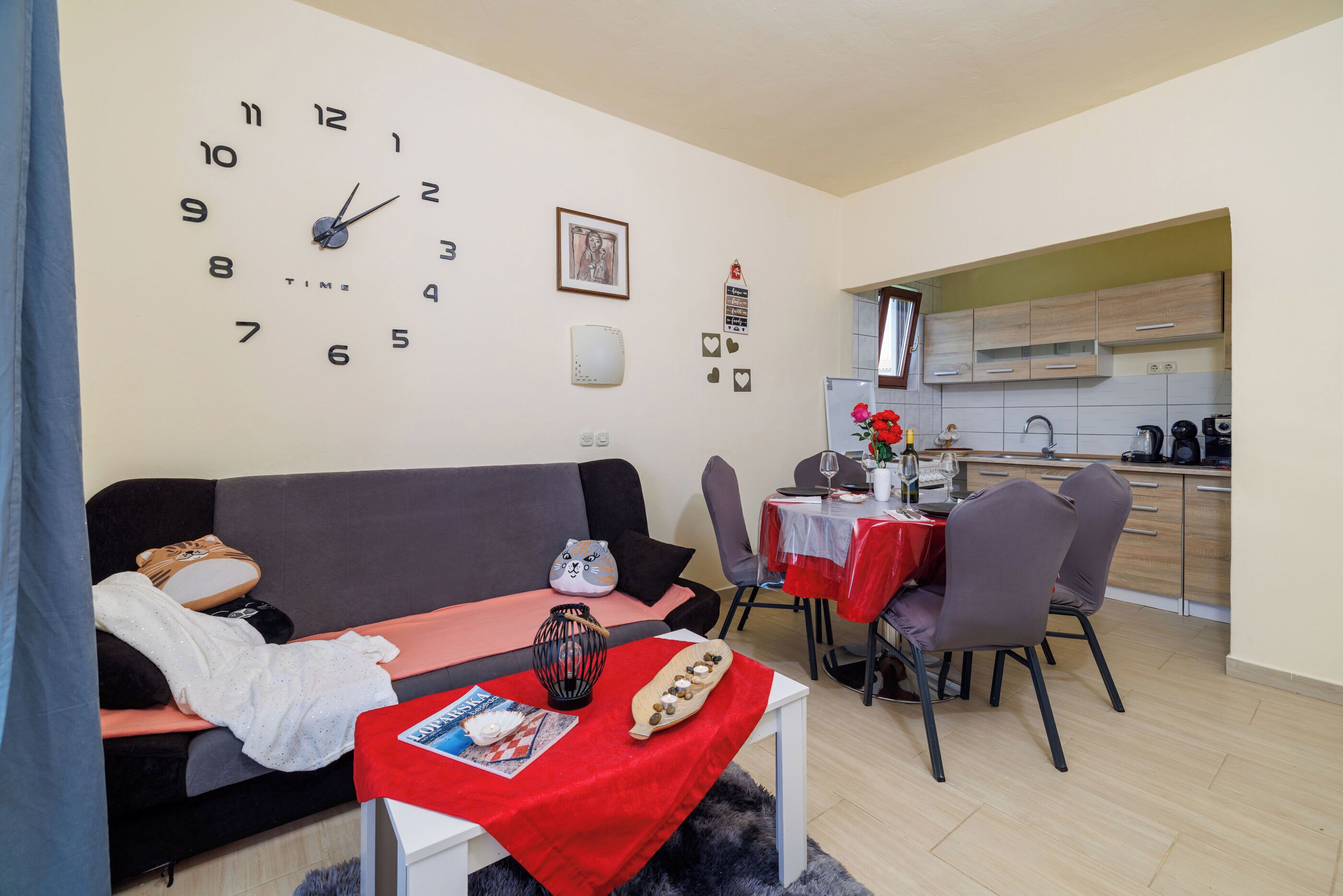 A modern living room with a sofa, a dining table with chairs, a kitchen area, and a wall clock.