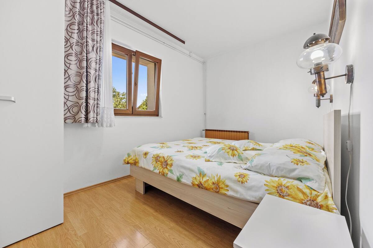 A bedroom with a wooden floor, a bed with a floral bedspread, a window with curtains, and a wall-mounted lamp.