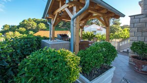 Eine hölzerne Pergola mit Grill und Sitzgelegenheit, umgeben von gepflegten Büschen und Kübelpflanzen.