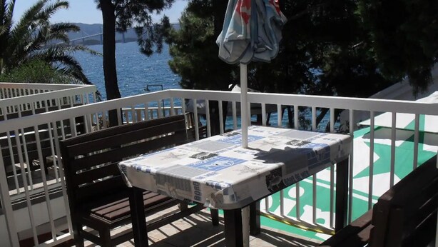 Ένα μπαλκόνι με ένα τραπέζι καλυμμένο με μια εφημερίδα, δύο ξύλινα παγκάκια και θέα στη θάλασσα και μια γέφυρα.