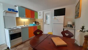 Μια μικρή, λειτουργική κουζίνα με εστία, νεροχύτη και πολύχρωμα ντουλάπια. Υπάρχει ένα τραπέζι φαγητού με καρέκλες και μια τηλεόραση επίπεδης οθόνης τοποθετημένη πάνω από το τραπέζι.