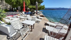 Ένας εξωτερικός χώρος με καθιστικά, λευκές ξαπλώστρες και τραπέζια, έναν πέτρινο τοίχο και θέα στη θάλασσα.