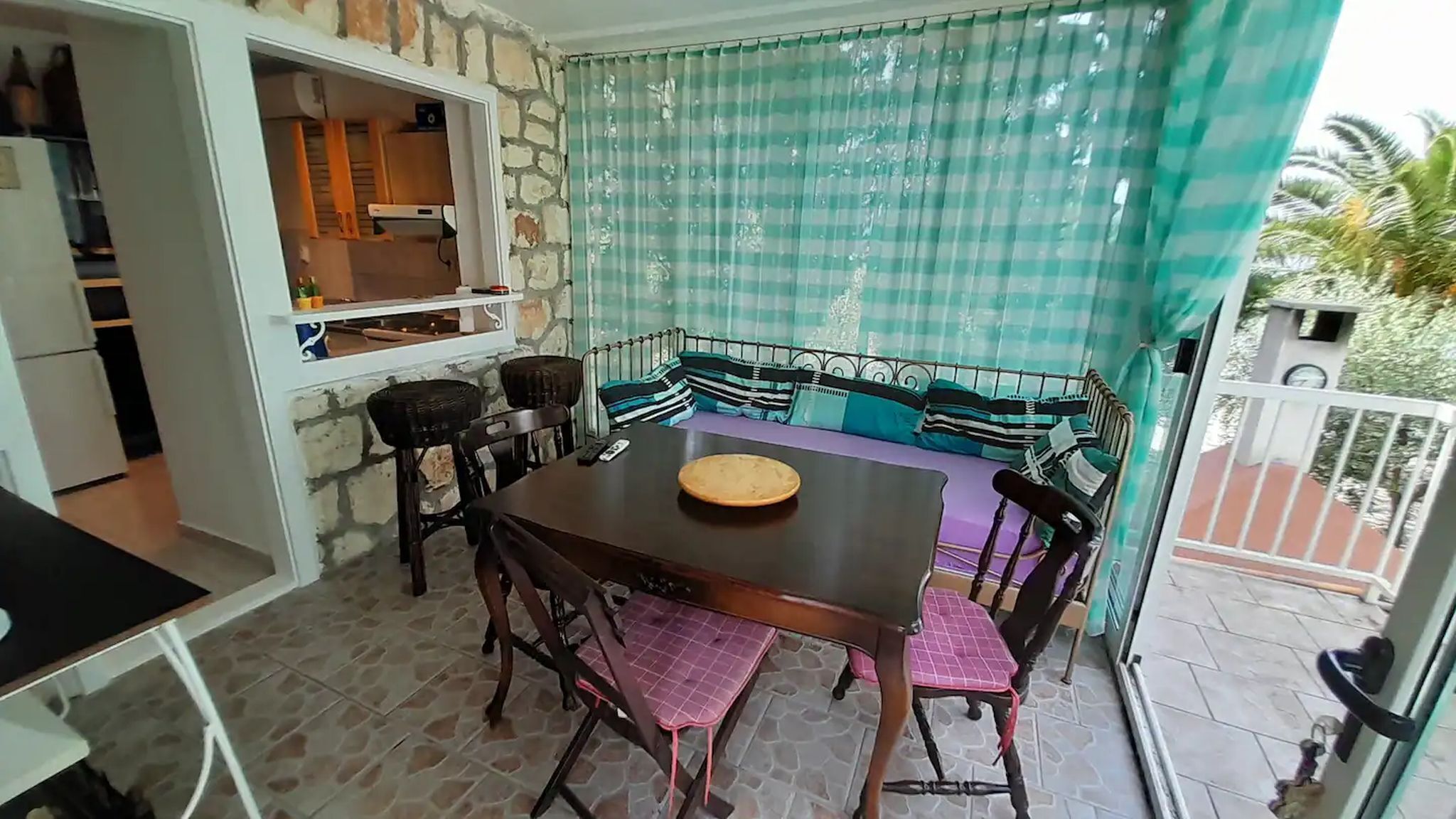 Et spiseområde med et bord og stole, et køkkenområde med et køleskab og hylder, samt en balkon med gardin i skakmønster.
