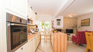 A modern kitchen with a built-in oven, a dining area with a wooden table and chairs, and a living room with a sofa and a TV. - House Srecko (Polaca)