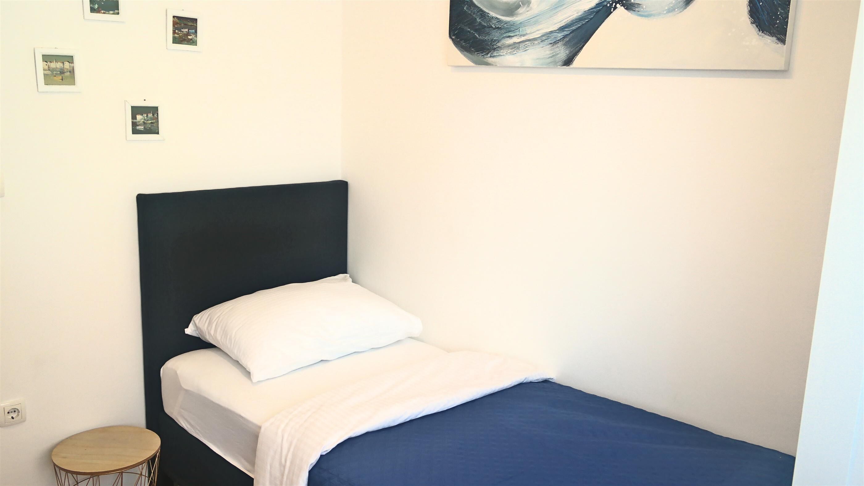 Una cama individual con cabecera oscura, ropa de cama blanca y una manta azul. Hay una mesita redonda pequeña al lado de la cama y un enchufe en la pared.