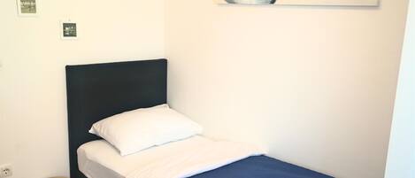Una cama individual con cabecera oscura, ropa de cama blanca y una manta azul. Hay una mesita redonda pequeña al lado de la cama y un enchufe en la pared.