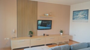A modern living room with a flat-screen TV mounted on the wall, a wooden cabinet with drawers, a glass coffee table, and a blue sofa. - Apartment Sarah (Cavle)