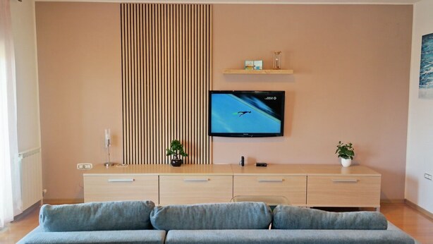 A modern living room with a flat-screen TV mounted on the wall, a wooden cabinet with drawers, a grey sofa, and a small potted plant. - Apartment Sarah (Cavle)
