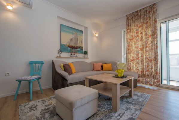 Una sala de estar moderna con un sofá, un reposapiés y una mesa de centro. Hay un cuadro en la pared, una silla y una ventana con cortinas.