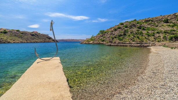 Una spiaggia di ciottoli che si estende verso acque calme, con una scultura in metallo a sinistra e un pendio roccioso a destra.