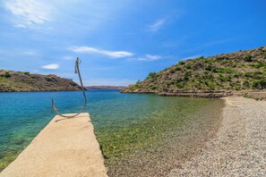 A clear view of a pebble beach leading into calm waters, with a metal sculpture on the left and a rocky hillside on the right. - Apartments Sanja (Karlobag)