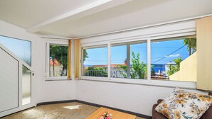 A room with a stone floor, a wooden table with a vase of flowers, a brown sofa with a floral cushion, and a view of a coastal area through large windows. - Apartment Car (Crikvenica)