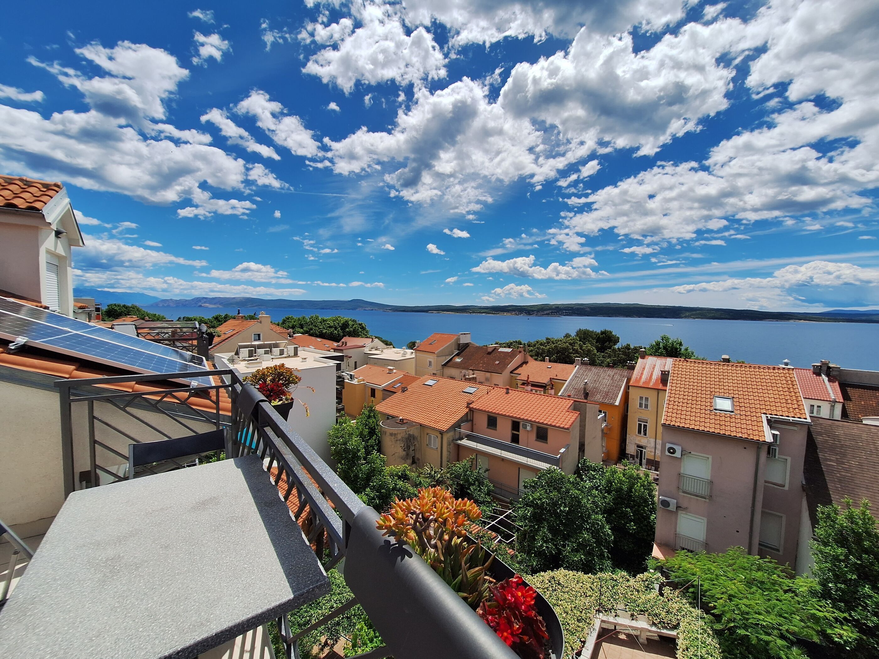 A residential area with terracotta-roofed buildings, a balcony with solar panels, and a view of a lake and mountains in the distance.